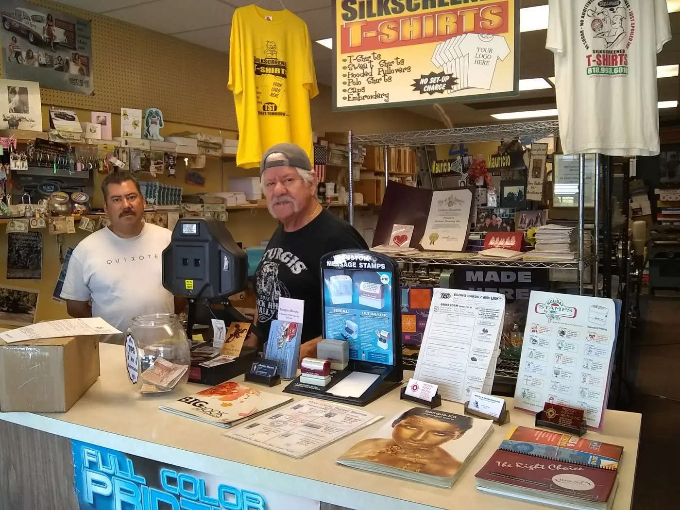 Mauricio and a colleague behind the counter at Precision Instant Printing, surrounded by sample cards, stamps and silkscreened shirts.